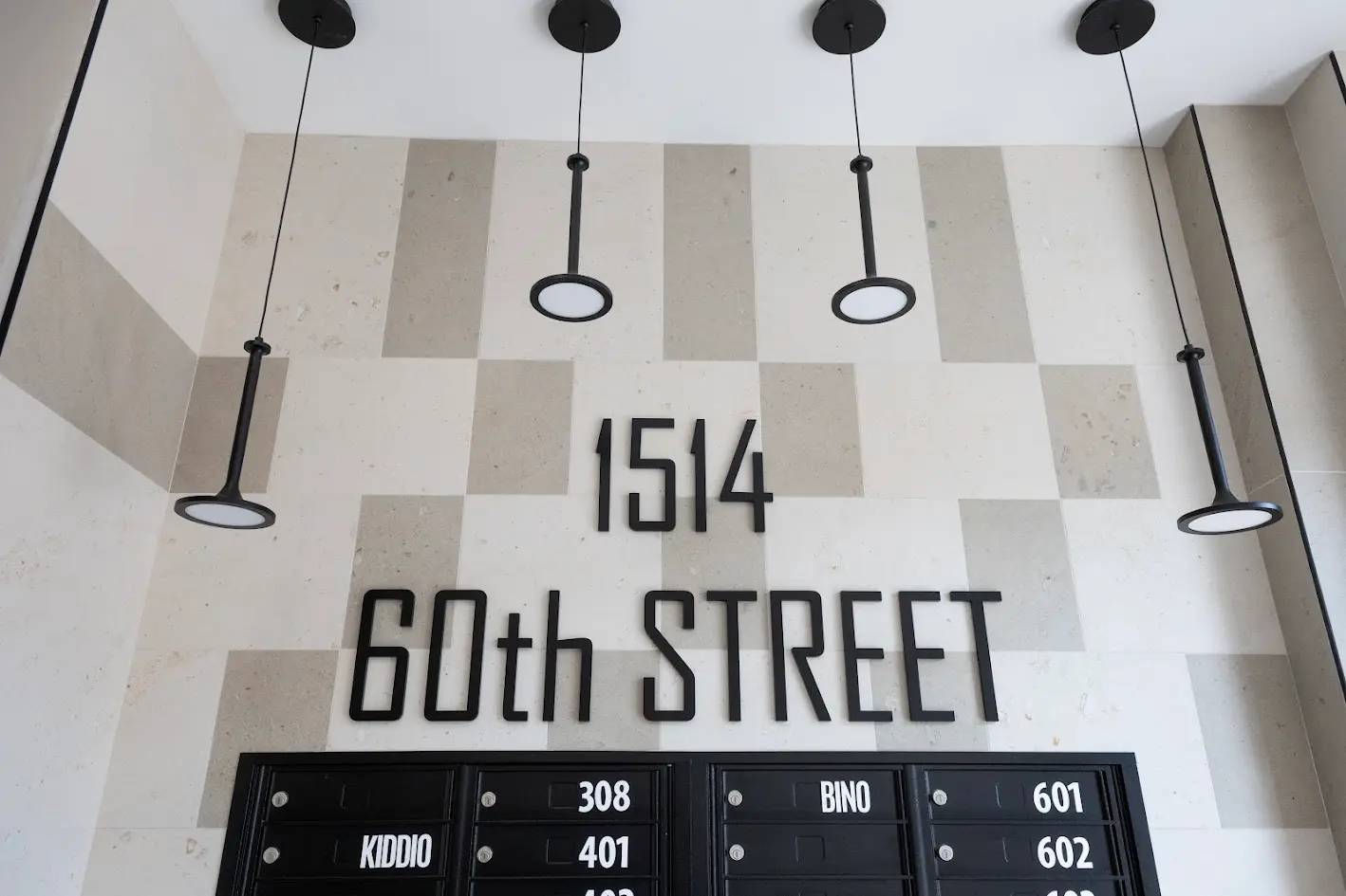 A modern building entrance with a checkered beige wall, black address numbers '1514 60th STREET', black mailboxes, and four black pendant lights.