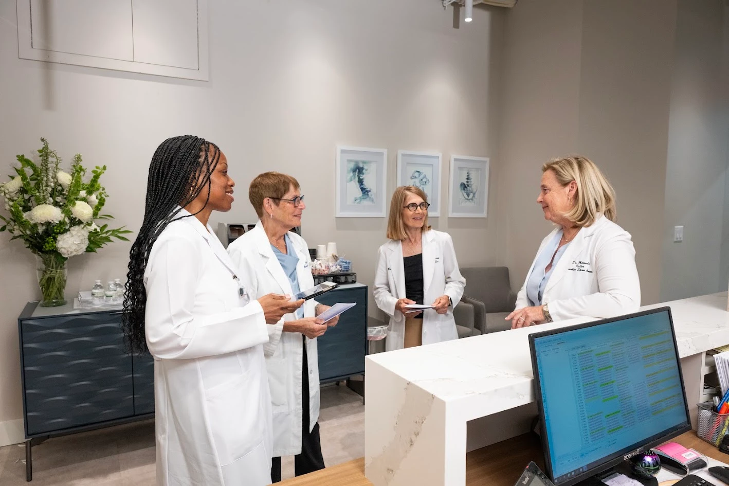 Four women in white lab coats smiling and talking in a modern medical office reception area.