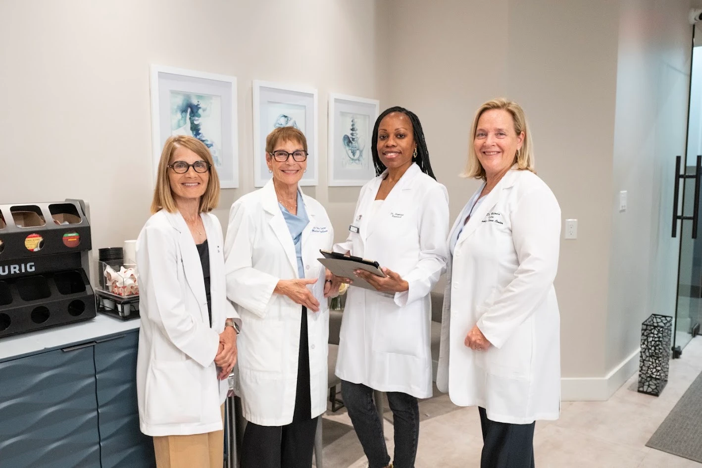 Four women in white lab coats stand smiling in a modern office setting with a Keurig station and framed artwork in the background.