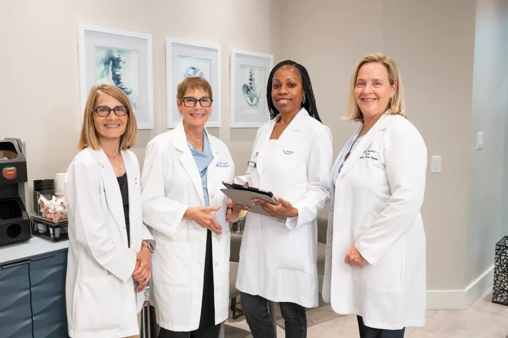 Four smiling women in white lab coats stand in a modern medical office, with anatomical artwork on the wall behind them.