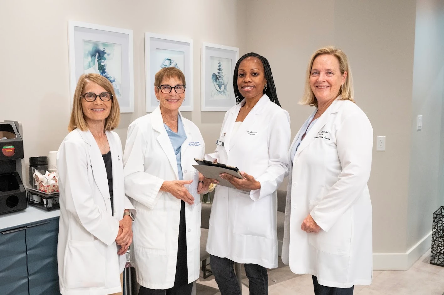 Four smiling women in white lab coats stand in a modern medical office, with anatomical artwork on the wall behind them.