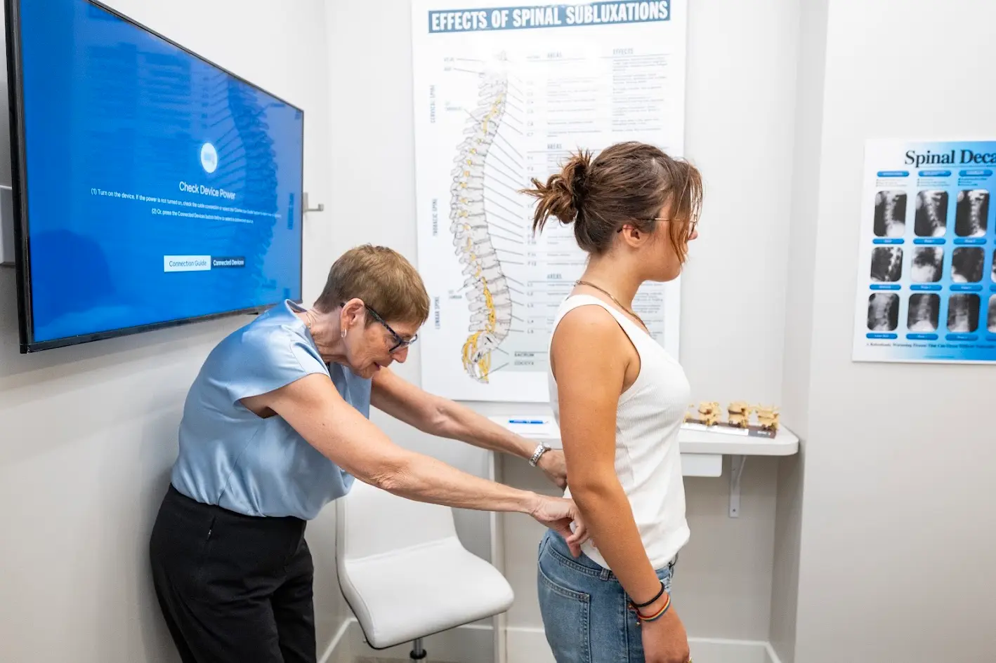 An older woman examines the lower back of a younger woman in a chiropractic office, with spinal health posters and a TV screen in the background.