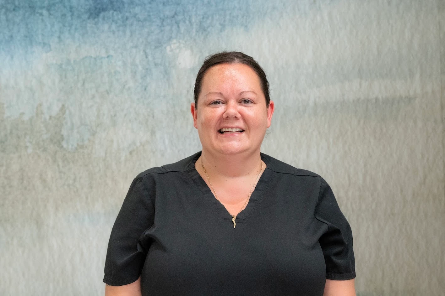 Headshot of a smiling woman with dark hair pulled back, wearing a black V-neck scrub top and a gold necklace, against a mottled blue and gray background.