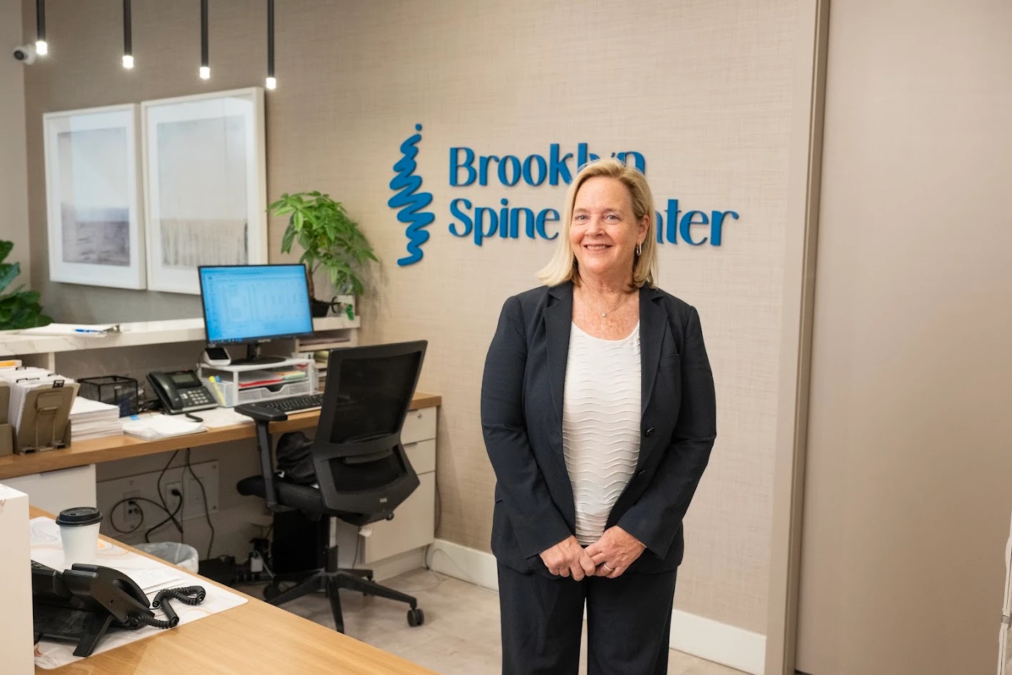 A smiling woman in a dark suit stands in the reception area of Brooklyn Spine Center, with a desk and computer to her left and the clinic's logo on the wall behind her.