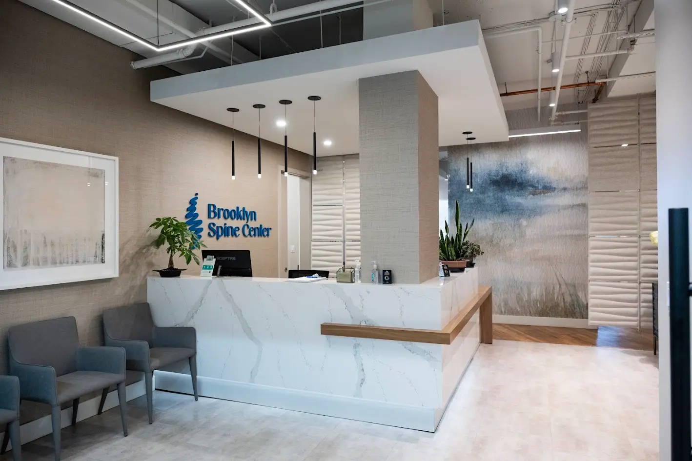A modern medical office reception area with a white marble-patterned L-shaped desk, gray waiting chairs, a 'Brooklyn Spine Center' logo, and abstract wall art including a landscape mural.
