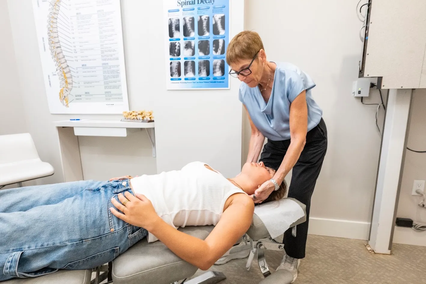 A chiropractor adjusts a patient's neck on a chiropractic table, with anatomical posters in the background.