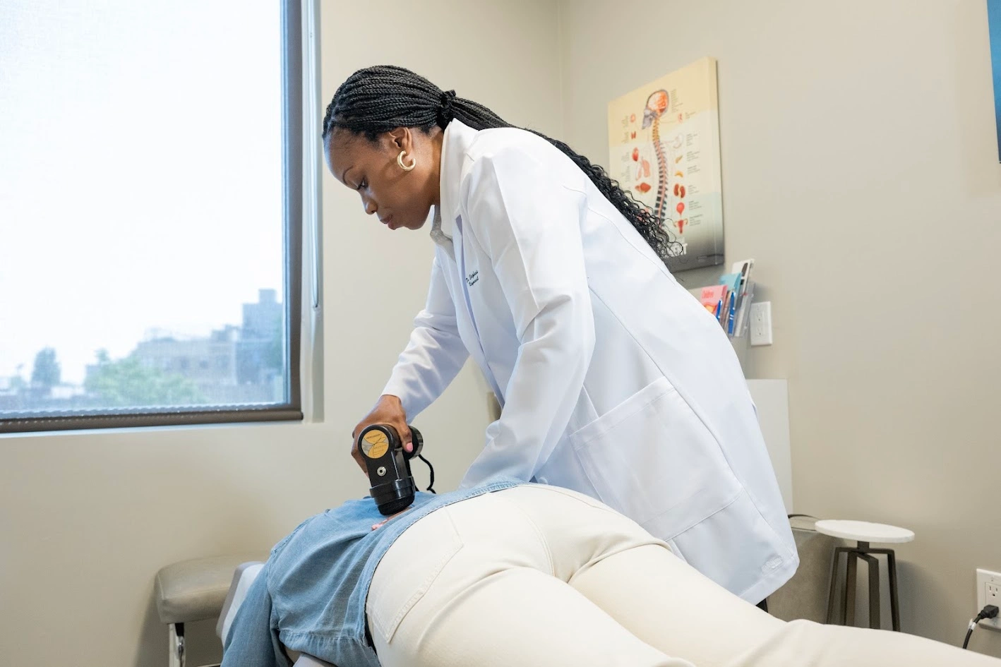 A medical professional applies a handheld device to a patient's lower back on an examination table.
