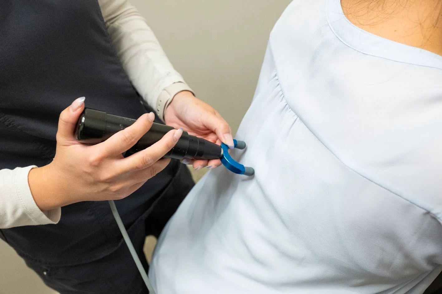 Close-up of a medical professional's hands holding a black device with a blue attachment, applying it to a patient's upper back.
