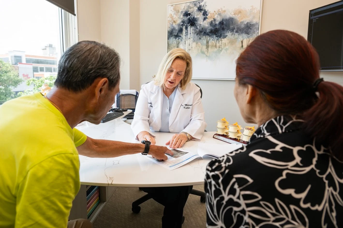 A female doctor in a white coat explains something to a male and female patient at a desk, pointing at a document. Spinal models are on the desk.