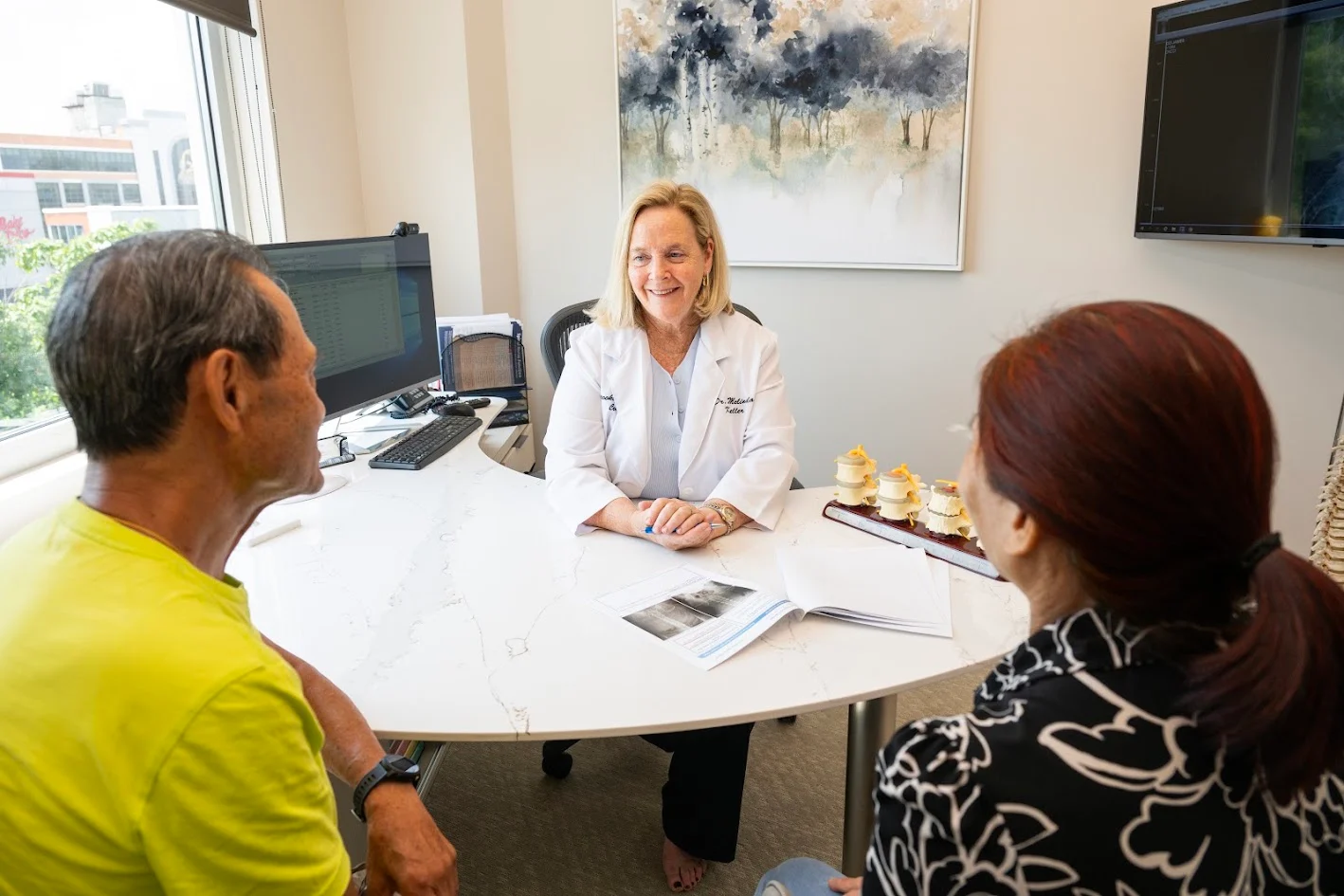 A smiling female doctor in a lab coat consults with an older male and female patient across a desk, with spinal models and medical images visible.