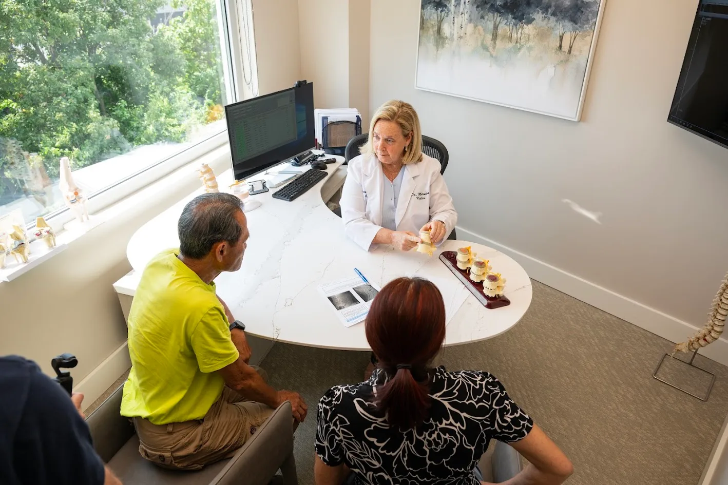 A female doctor in a white lab coat explains a spinal model to a male and female patient in a bright consultation room.