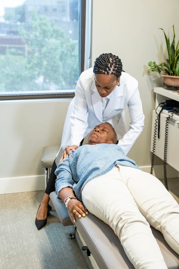 A Black female doctor in a white lab coat examines an older Black female patient lying on an examination table, placing her hands on the patient's shoulders.