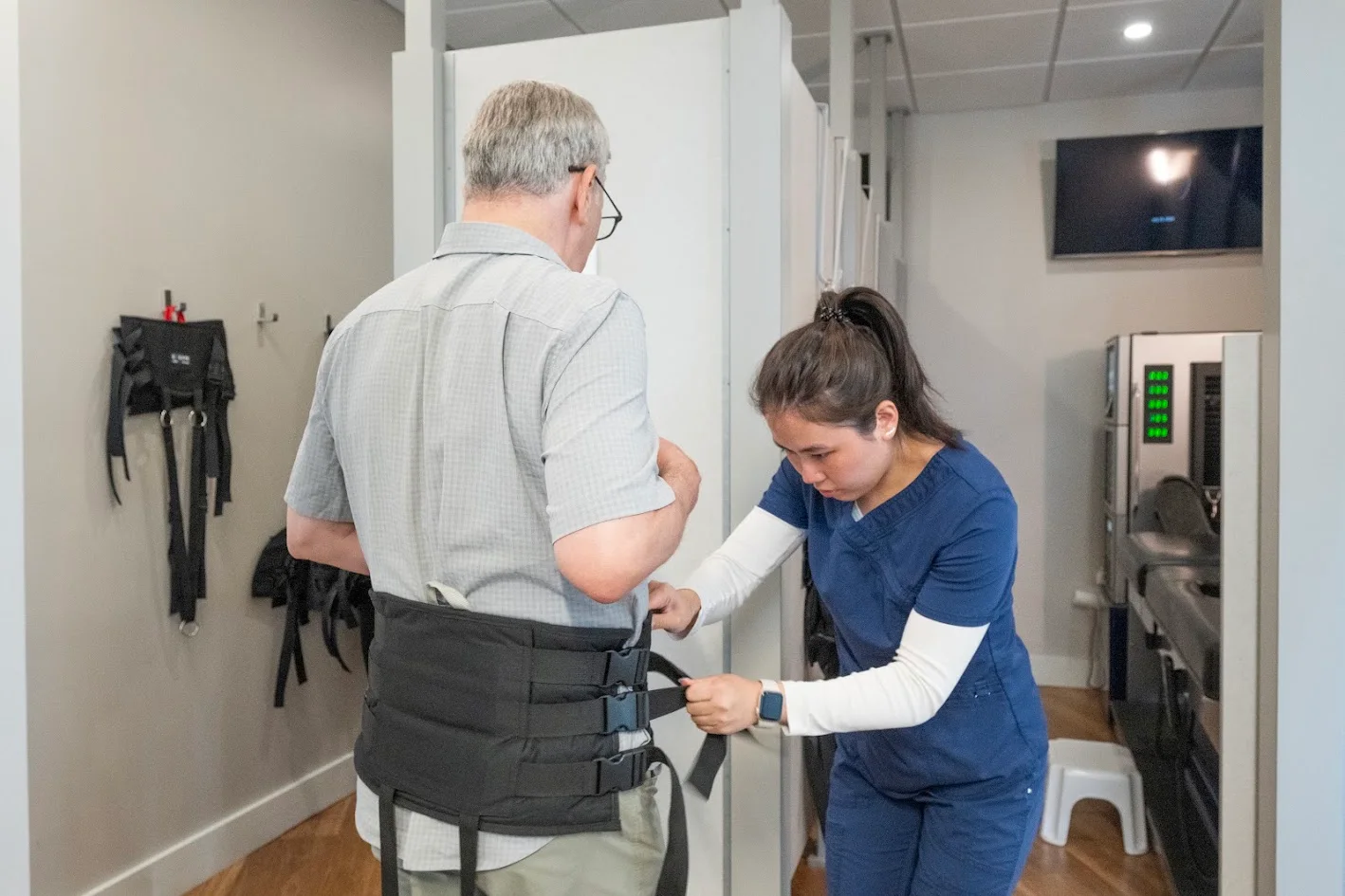 A woman in blue scrubs helps a man fit a black back brace around his lower back in a clinic setting.