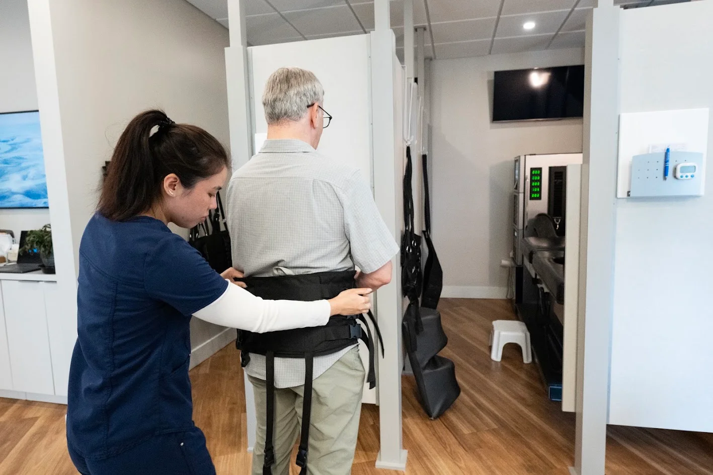A female therapist adjusts a black harness on a male patient's waist in a clinic.