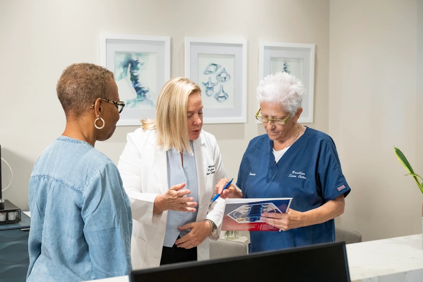 Three women, two in medical attire, discussing a brochure at a reception desk in a medical office.