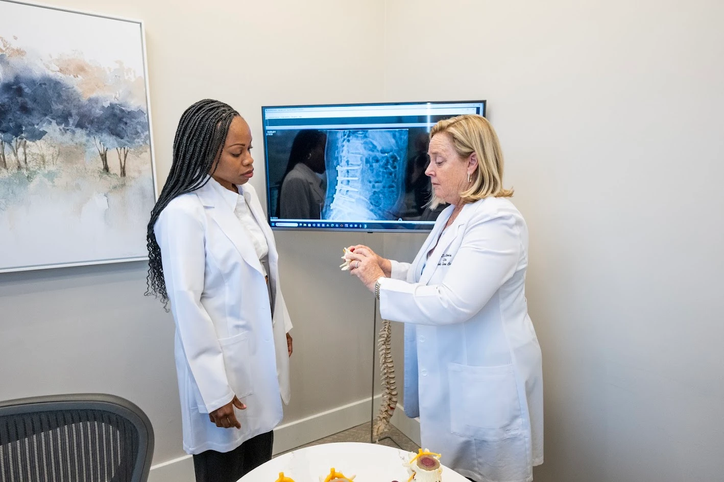 Two women in white lab coats, one explaining a spinal model while the other listens, with a spinal X-ray on a screen in the background.