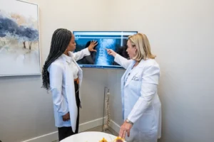 Two doctors in white lab coats examining a spinal X-ray on a monitor, with a spine model and disc models on a table.
