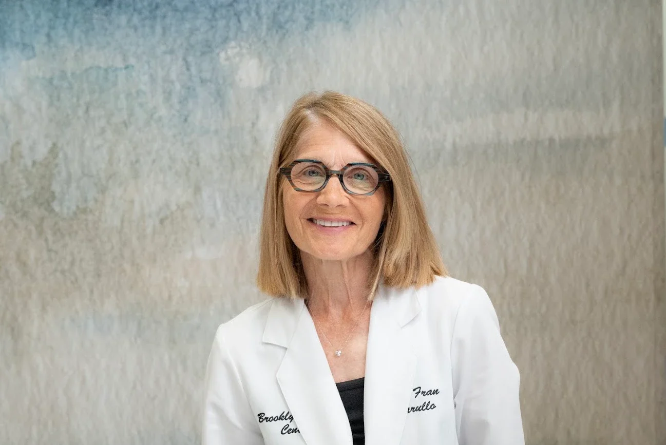 A smiling woman with blonde hair and glasses in a white lab coat, with 'Brooklyn Cen' and 'Fran Sarullo' embroidered on it, stands against a muted watercolor wall.