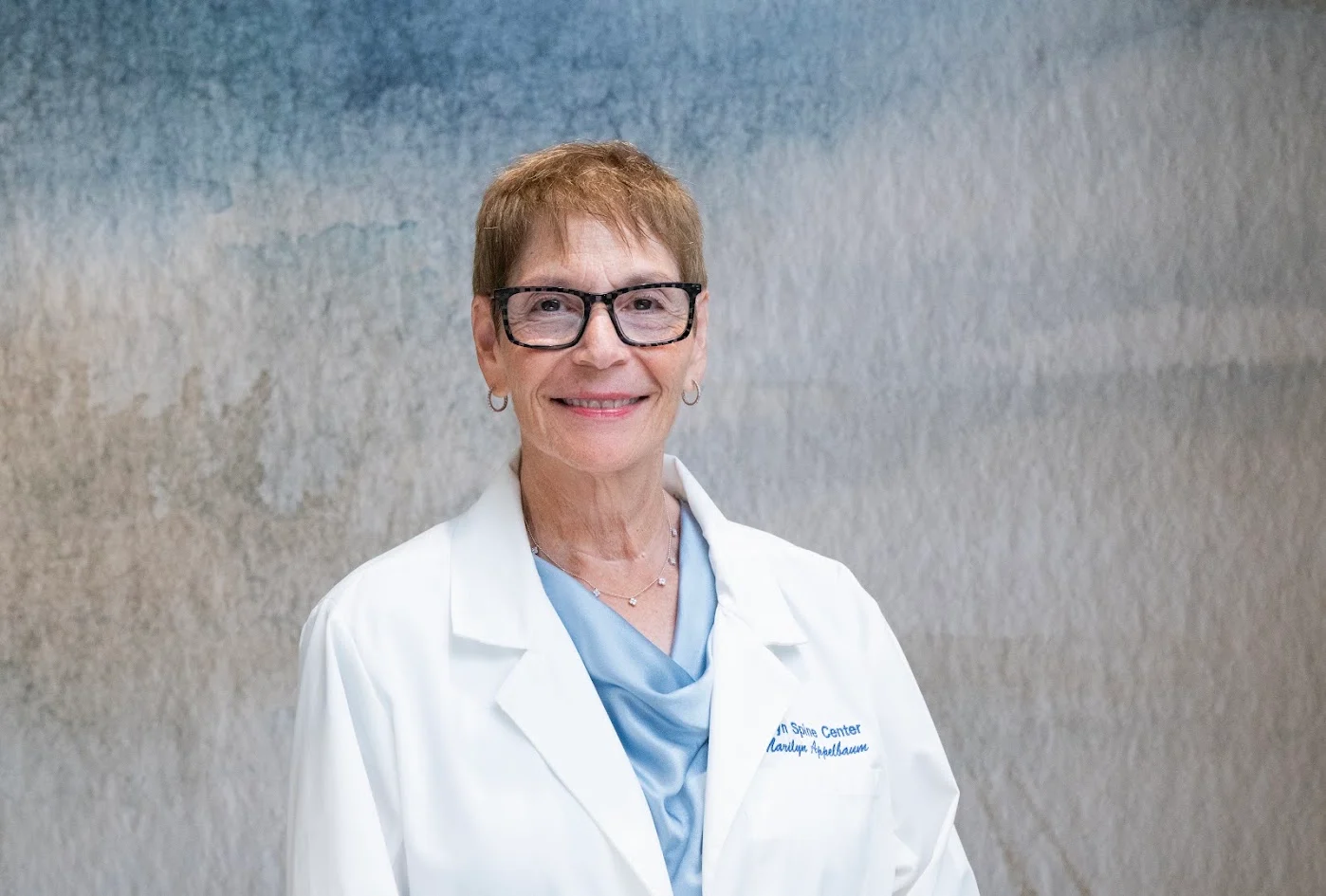 Headshot of Marilyn Appelbaum, a woman with short hair and glasses, wearing a white lab coat over a blue top, smiling.
