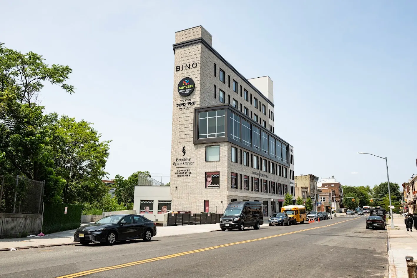 A tall, narrow, light-brick commercial building with multiple businesses, including Brooklyn Spine Center, on an urban street with parked cars and trees.