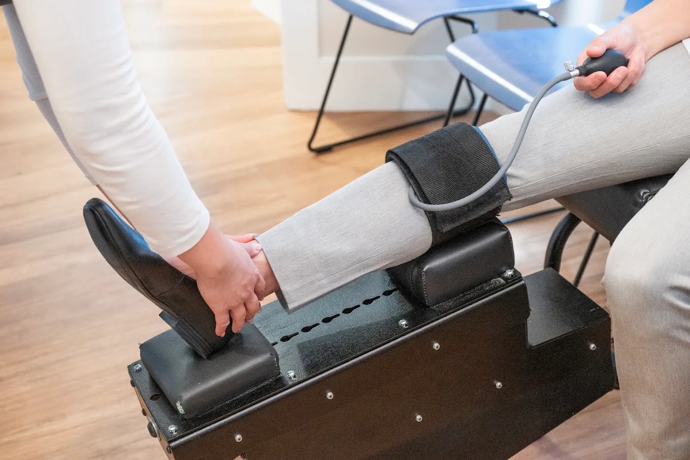 A medical professional holds a patient's foot while their leg rests on a black medical device with a blood pressure cuff on the calf. The patient holds the cuff's inflation bulb.