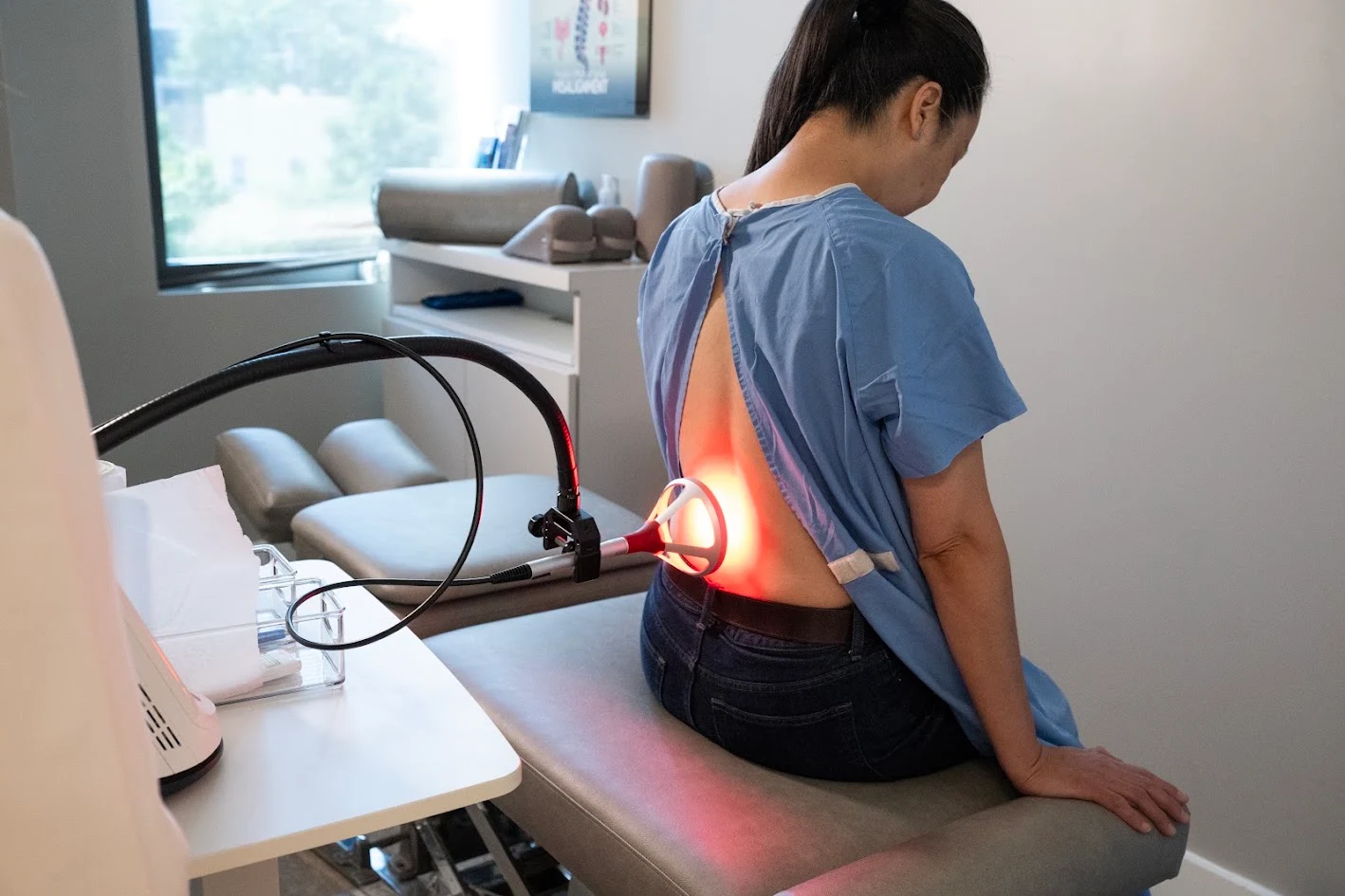A person in a medical gown sits on an examination table receiving red light therapy on their lower back.