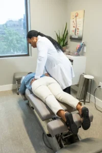 A chiropractor in a white lab coat adjusts a patient's lower back on an examination table in a clinic.