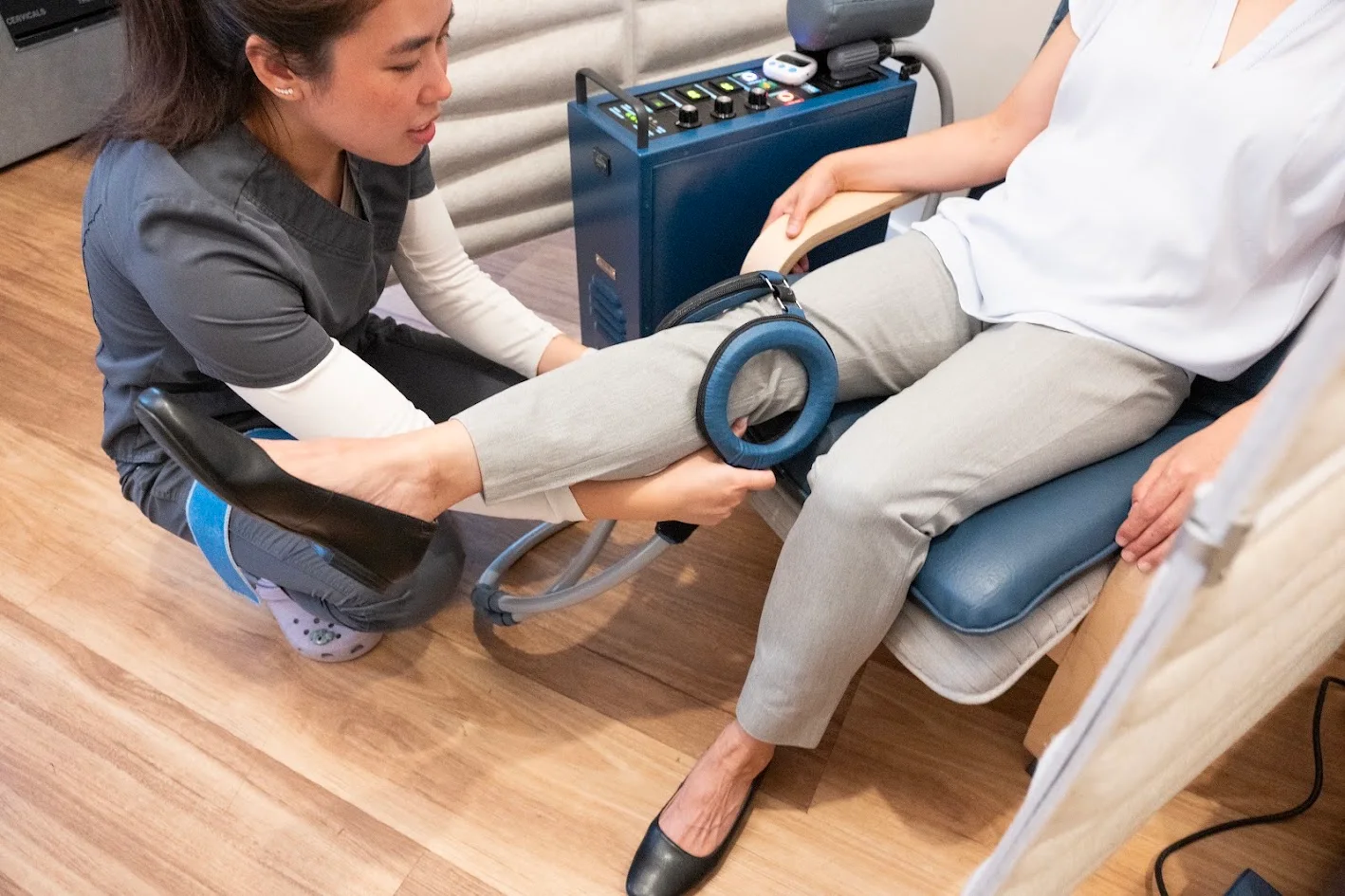 A medical professional in scrubs kneels, applying a blue circular device to a patient's knee while the patient sits in a chair.