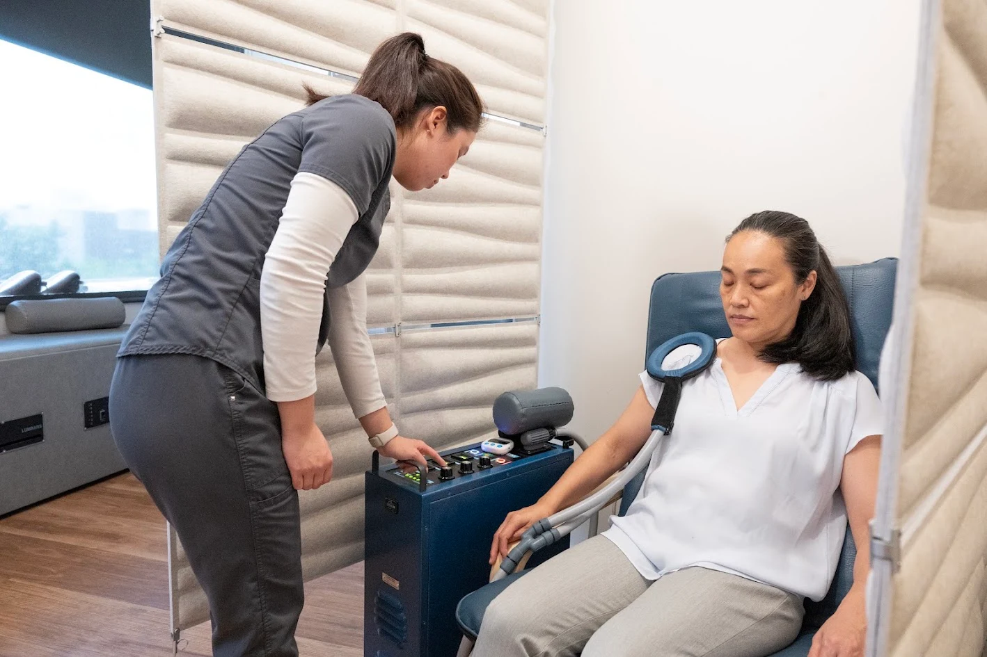 A healthcare professional adjusts a medical machine while a patient sits in a chair with a device on her shoulder, eyes closed.