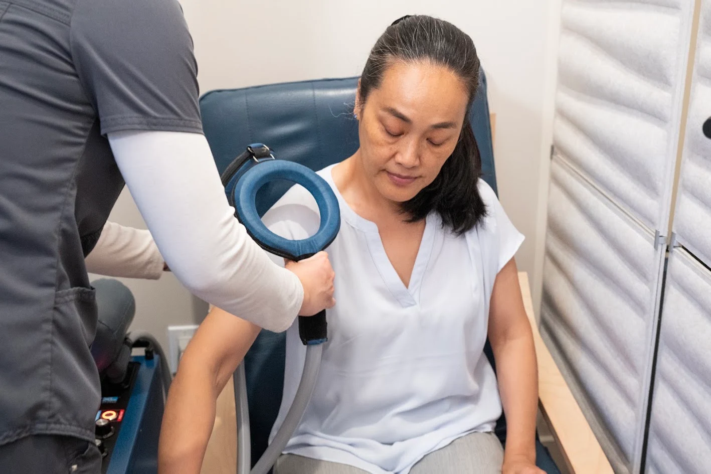 A woman sits in a chair while a medical professional applies a blue, circular device to her upper arm.