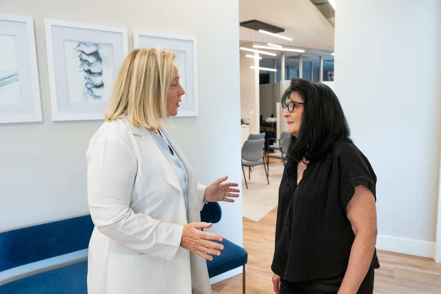 Two women, one in a white lab coat and one in a black top, stand and talk in a bright, modern clinic.
