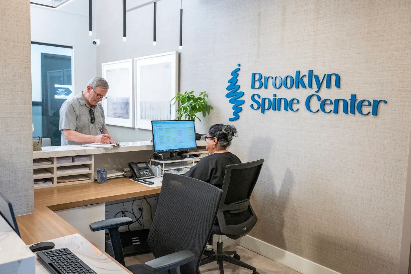 A man writes on a clipboard at the reception counter of Brooklyn Spine Center, while a woman works at a computer behind the desk.