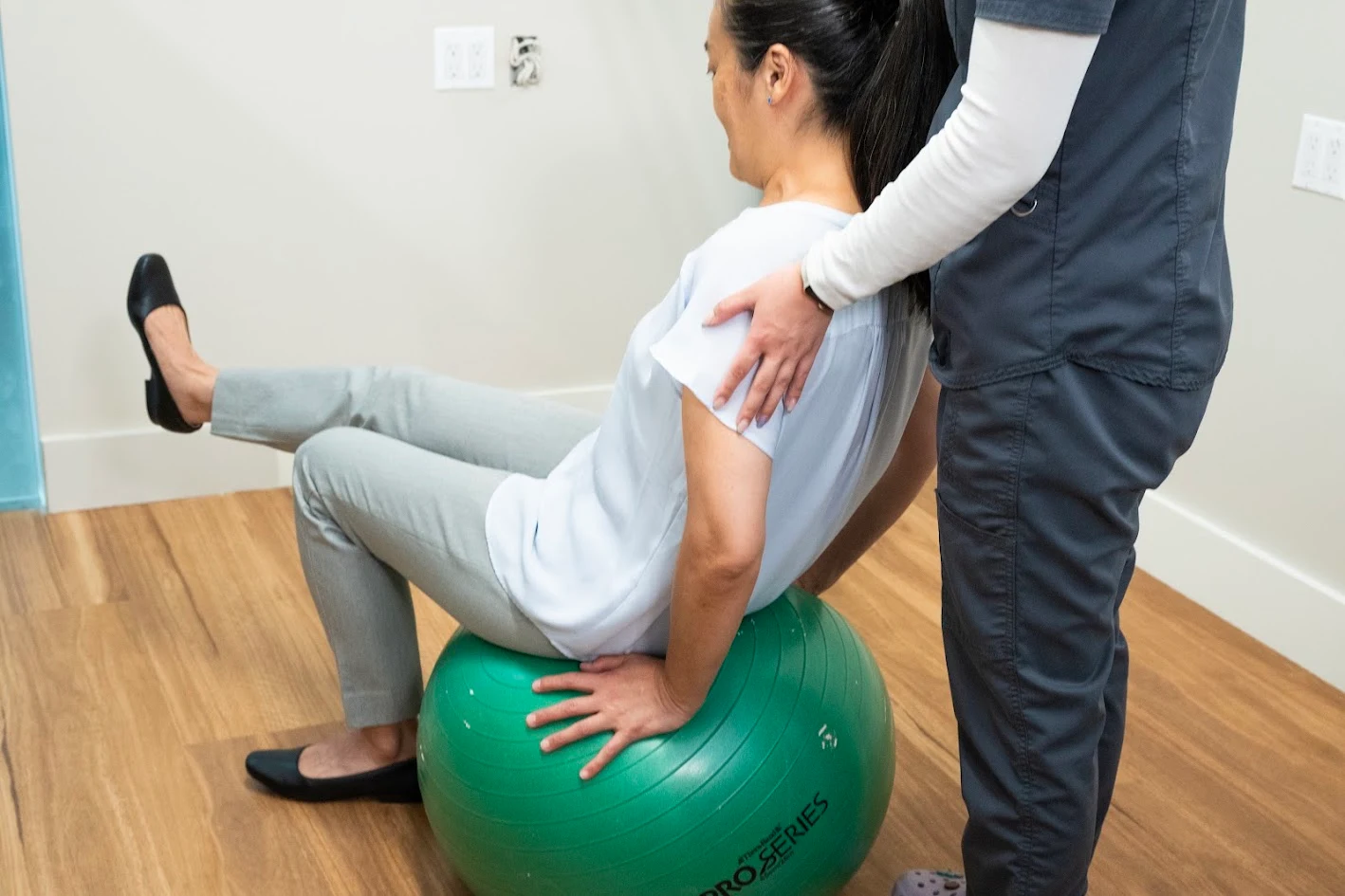 A person sitting on a green exercise ball with one leg extended, supported by a professional standing behind them.