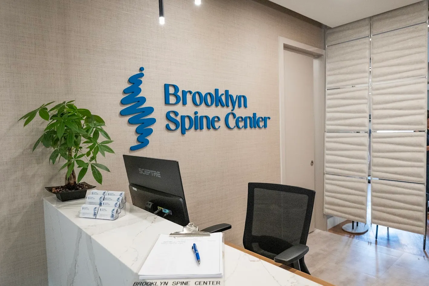 Reception desk at Brooklyn Spine Center with a blue logo, monitor, plant, and office chair.
