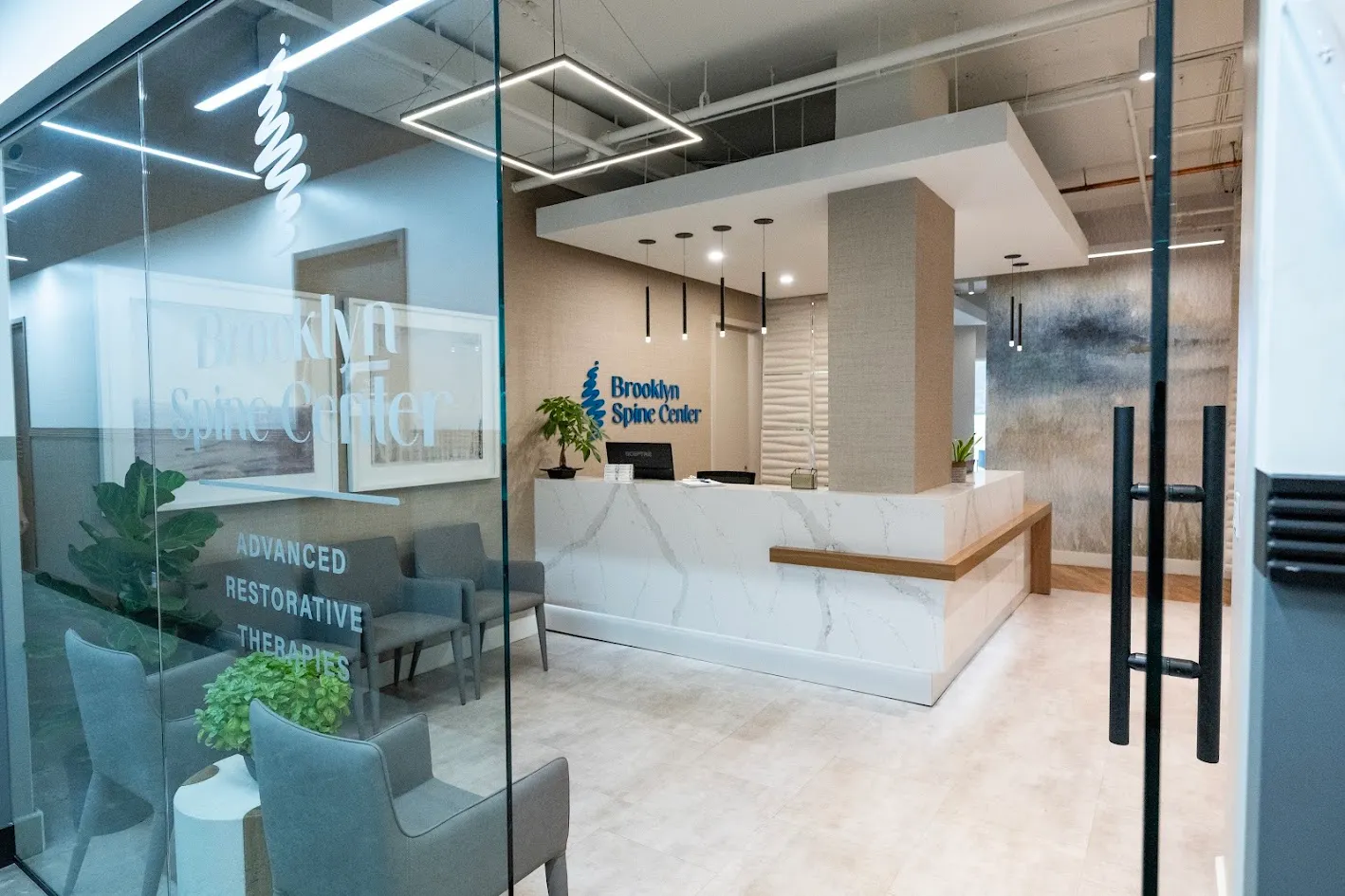 A modern and clean reception area of the Brooklyn Spine Center, featuring a white marble-patterned reception desk, grey armchairs, and a glass partition with the clinic's name.