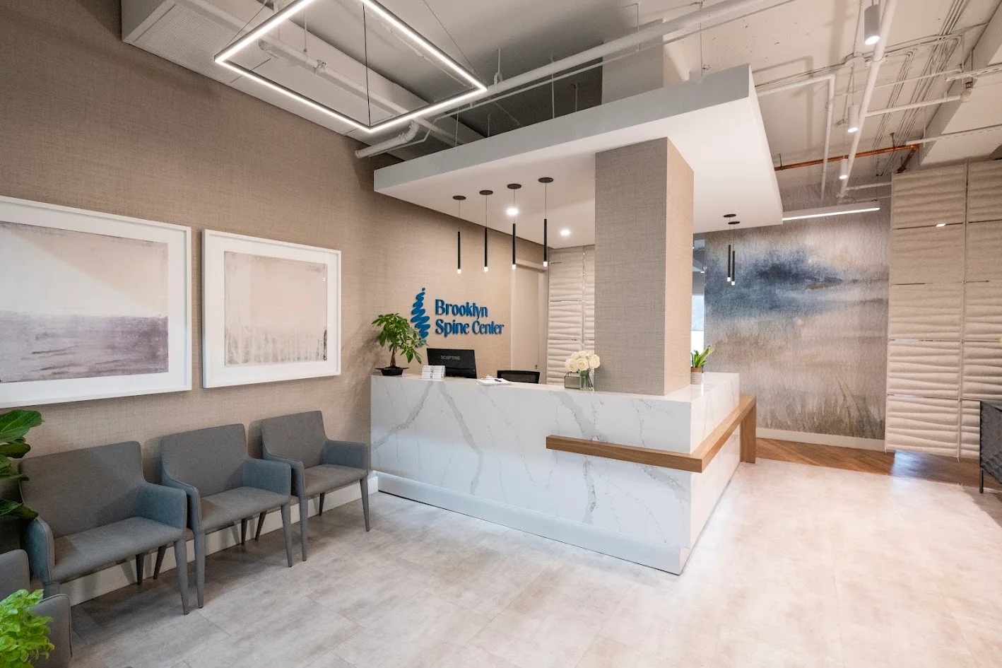 A modern reception area featuring a white marble desk with the 'Brooklyn Spine Center' logo, gray waiting chairs, and abstract art on textured beige walls.