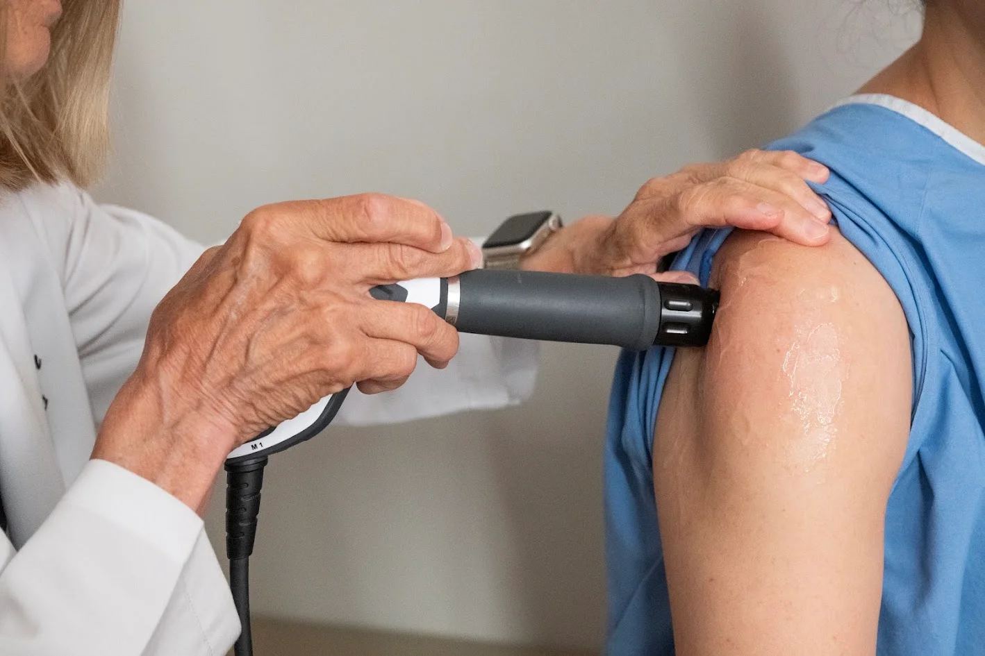 Close-up of a medical professional's hands holding a device to a patient's gel-covered shoulder.
