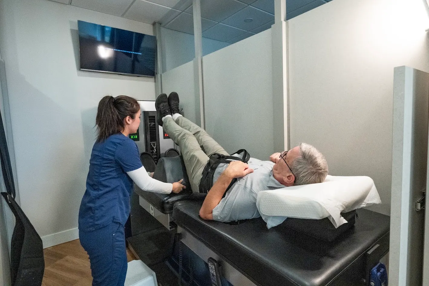 A medical professional in blue scrubs assists an older man lying on his back on a therapy machine with his legs elevated.