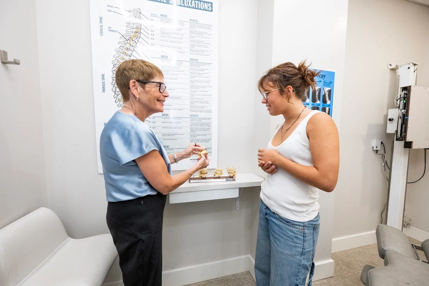 An older female chiropractor explains spinal models to a young female patient in a medical office with a spine chart on the wall.