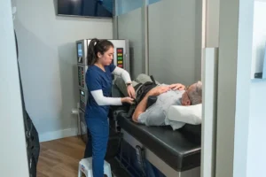 A female medical professional in blue scrubs adjusts a harness on an older male patient lying on a black medical table.