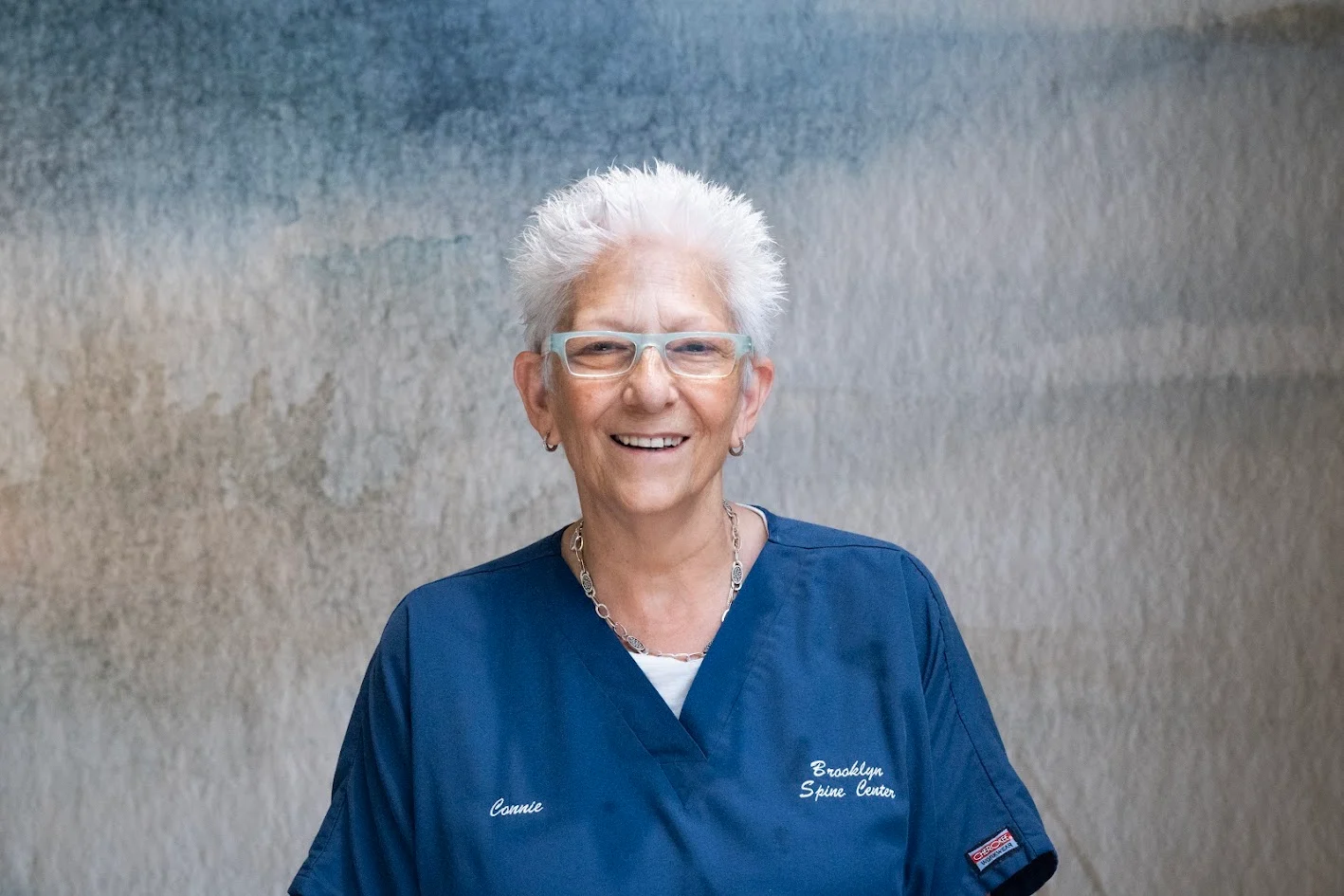 Headshot of a smiling woman with white spiky hair and glasses, wearing blue scrubs with 'Connie' and 'Brooklyn Spine Center' embroidered on them.