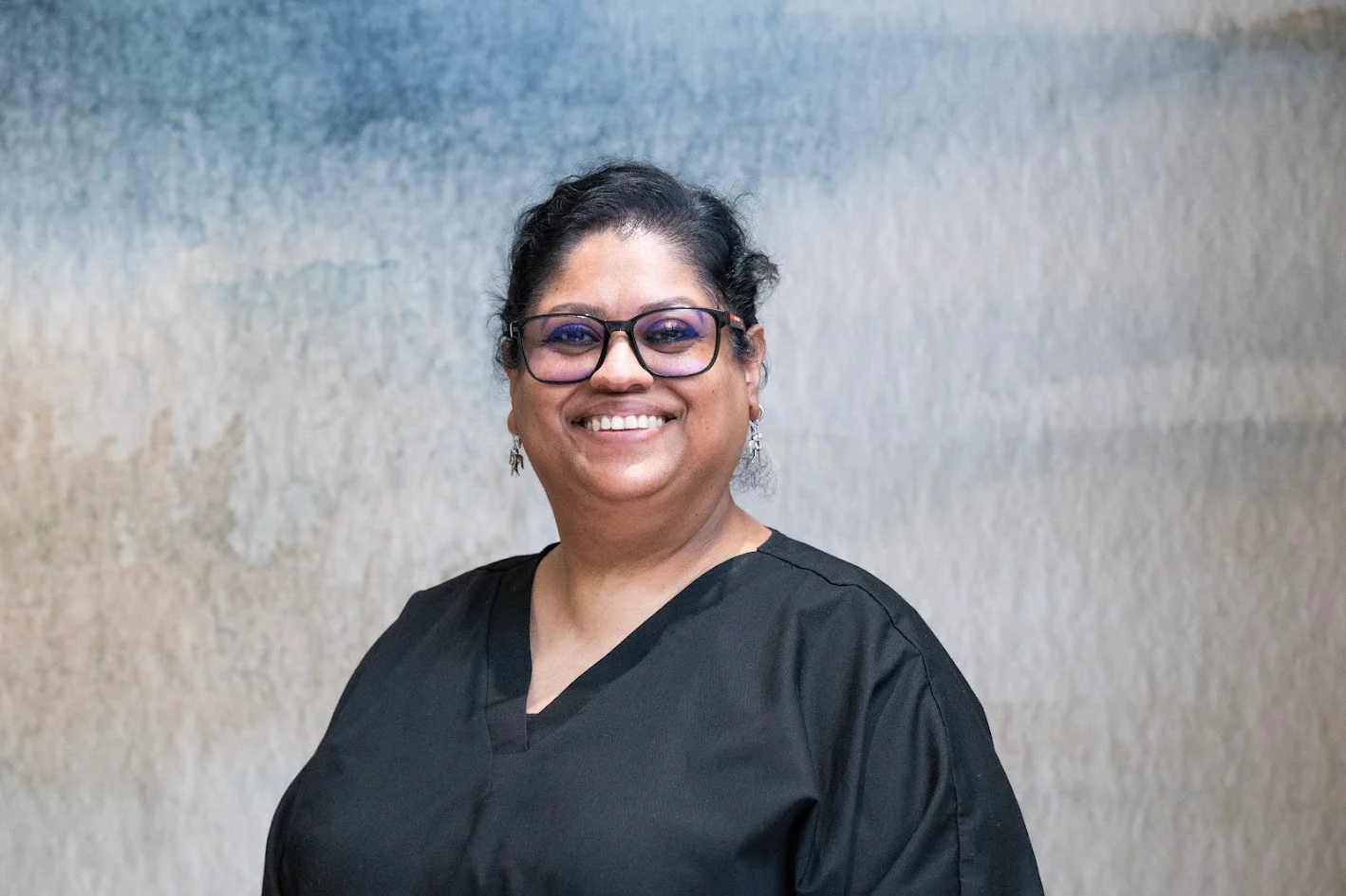 Headshot of a smiling woman with dark, curly hair, wearing glasses and a black scrub top against a blurred blue and gray background.